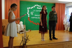 Uczniowie na scenie śpiewają pieśni. Koło sceny poczet sztandarowy, na scenie uczniowie przebrani w czarne stroje zaborców i  na biało za orła. Uczniowie składają znicze pod krzyżem.