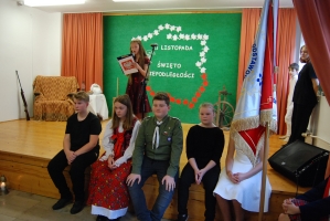 Uczniowie na scenie śpiewają pieśni. Koło sceny poczet sztandarowy, na scenie uczniowie przebrani w czarne stroje zaborców i  na biało za orła. Uczniowie składają znicze pod krzyżem.