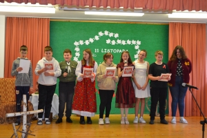 Uczniowie na scenie śpiewają pieśni. Koło sceny poczet sztandarowy, na scenie uczniowie przebrani w czarne stroje zaborców i  na biało za orła. Uczniowie składają znicze pod krzyżem.