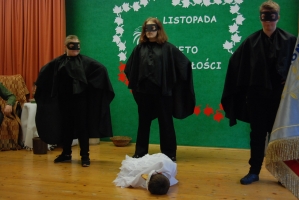 Uczniowie na scenie śpiewają pieśni. Koło sceny poczet sztandarowy, na scenie uczniowie przebrani w czarne stroje zaborców i  na biało za orła. Uczniowie składają znicze pod krzyżem.