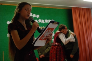 Uczniowie na scenie śpiewają pieśni. Koło sceny poczet sztandarowy, na scenie uczniowie przebrani w czarne stroje zaborców i  na biało za orła. Uczniowie składają znicze pod krzyżem.