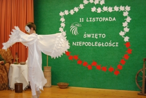 Uczniowie na scenie śpiewają pieśni. Koło sceny poczet sztandarowy, na scenie uczniowie przebrani w czarne stroje zaborców i  na biało za orła. Uczniowie składają znicze pod krzyżem.