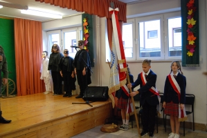 Uczniowie na scenie śpiewają pieśni. Koło sceny poczet sztandarowy, na scenie uczniowie przebrani w czarne stroje zaborców i  na biało za orła. Uczniowie składają znicze pod krzyżem.