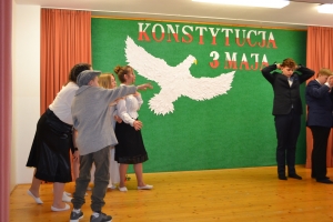 Uczniowie na scenie. Przebrani w stroje biało czarne i ułańskie z epoki. Tańczą, markują ruchy kopania, ogrzewania przy ognisku.