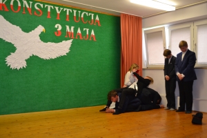 Uczniowie na scenie. Przebrani w stroje biało czarne i ułańskie z epoki. Tańczą, markują ruchy kopania, ogrzewania przy ognisku.