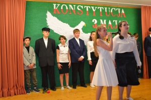 Uczniowie na scenie. Przebrani w stroje biało czarne i ułańskie z epoki. Tańczą, markują ruchy kopania, ogrzewania przy ognisku.