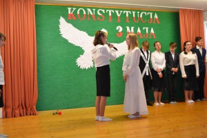 Uczniowie na scenie. Przebrani w stroje biało czarne i ułańskie z epoki. Tańczą, markują ruchy kopania, ogrzewania przy ognisku.