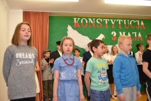 Uczniowie na scenie. Przebrani w stroje biało czarne i ułańskie z epoki. Tańczą, markują ruchy kopania, ogrzewania przy ognisku.