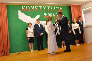 Uczniowie na scenie. Przebrani w stroje biało czarne i ułańskie z epoki. Tańczą, markują ruchy kopania, ogrzewania przy ognisku.