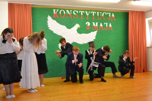 Uczniowie na scenie. Przebrani w stroje biało czarne i ułańskie z epoki. Tańczą, markują ruchy kopania, ogrzewania przy ognisku.