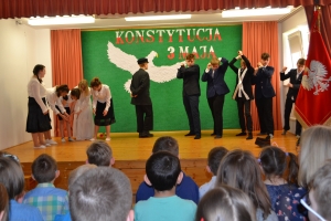 Uczniowie na scenie. Przebrani w stroje biało czarne i ułańskie z epoki. Tańczą, markują ruchy kopania, ogrzewania przy ognisku.