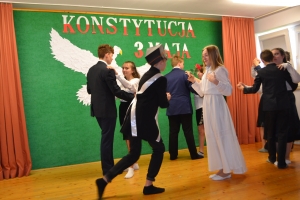 Uczniowie na scenie. Przebrani w stroje biało czarne i ułańskie z epoki. Tańczą, markują ruchy kopania, ogrzewania przy ognisku.