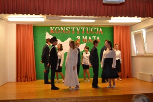 Uczniowie na scenie. Przebrani w stroje biało czarne i ułańskie z epoki. Tańczą, markują ruchy kopania, ogrzewania przy ognisku.