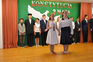 Uczniowie na scenie. Przebrani w stroje biało czarne i ułańskie z epoki. Tańczą, markują ruchy kopania, ogrzewania przy ognisku.