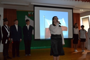 Uczniowie na scenie. Przebrani w stroje biało czarne i ułańskie z epoki. Tańczą, markują ruchy kopania, ogrzewania przy ognisku.