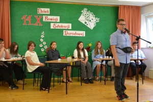 Uczniowie występują na scenie w galowych strojach, dyrektor pasuje uczniów długim ołówkiem