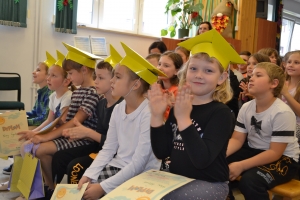 Uczniowie występują na scenie w galowych strojach, dyrektor pasuje uczniów długim ołówkiem