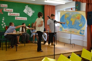 Uczniowie występują na scenie w galowych strojach, dyrektor pasuje uczniów długim ołówkiem