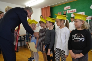 Uczniowie występują na scenie w galowych strojach, dyrektor pasuje uczniów długim ołówkiem