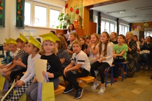 Uczniowie występują na scenie w galowych strojach, dyrektor pasuje uczniów długim ołówkiem