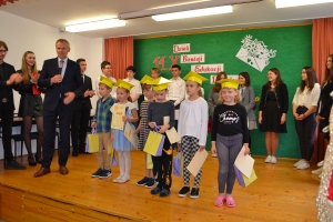 Uczniowie występują na scenie w galowych strojach, dyrektor pasuje uczniów długim ołówkiem