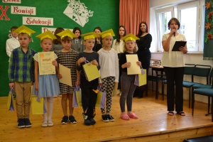 Uczniowie występują na scenie w galowych strojach, dyrektor pasuje uczniów długim ołówkiem