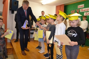 Uczniowie występują na scenie w galowych strojach, dyrektor pasuje uczniów długim ołówkiem