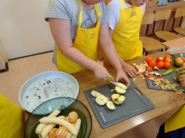 dzieci ubrane w fartuszki i czapki kucharskie, kroją warzywa